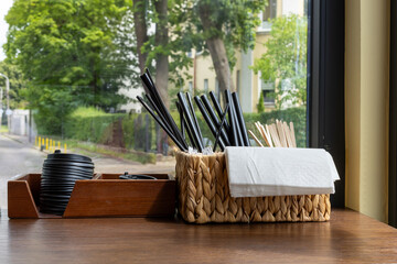 Set of black plastic straws, wooden stirrers, napkins and cup lids in containers on wooden counter...