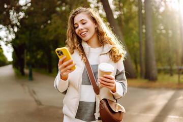 Obraz premium A smiling woman in casual clothes is walking in a sunny park with a phone and a cup of coffee. Young woman is chatting or blogging outdoors. Beauty, technology and walk concept.