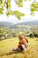 Hiker woman in hat sits on blanket on top of green hill enjoying sunny landscape. Beautiful woman feels freedom and walks in hills. Travel, leisure and freedom concept.