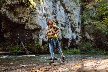 Young female traveler with hiking backpack near mountain river enjoys mountain landscape. Curly woman feels relaxed in nature and walks through forest in the mountains. Travel concept, activity.
