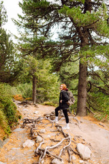 Obraz premium Female hiker with a backpack stands on mountain hiking trail and enjoys the mountain scenery on a sunny day. Young woman smiles and walks in forest. Concept of relaxation, nature. Active lifestyle.