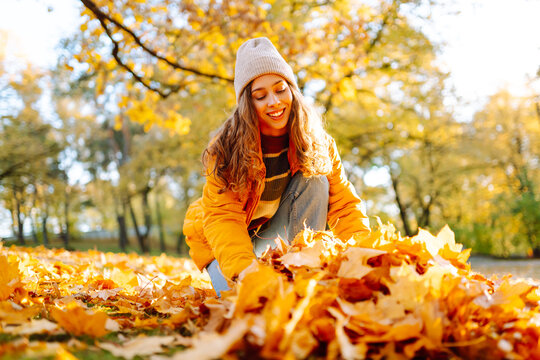 Close-up of a female volunteer in an orange jacket and gloves collects bright leaves on a sunny day. Young woman holds pile of autumn leaves in park. Concept of volunteering, seasonality and ecology.
