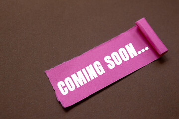 Coming soon message written on ripped torn pink paper piece on brown background. Conceptual coming soon symbol. Copy space.