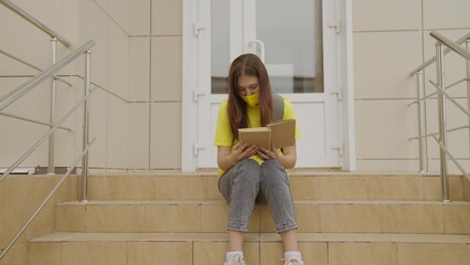 A schoolgirl girl in a mask reads a book while standing at the school, protection against...
