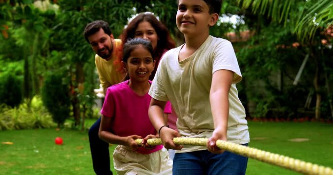 Indian family playing tug of war rassi khech in home garden, spending quality fun time together enjoying outdoor sports, cheerful Asian parents children bonding through traditional backyard activity
