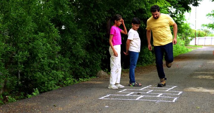 Indian family playing Tikkar Billa, a traditional hopscotch game in home garden, spending quality fun time together with children hopping on squares and enjoying nostalgic outdoor sports activity