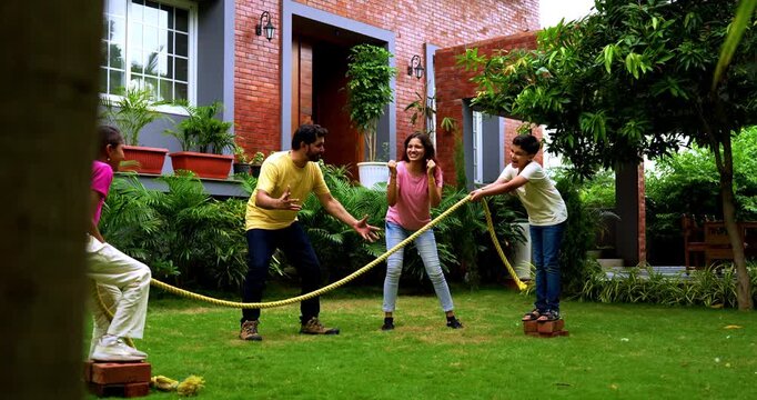 Indian kids playing tug of war or rassi khech in home garden while parents cheer them, spending quality fun time together enjoying traditional outdoor sports bonding during weekend family activity