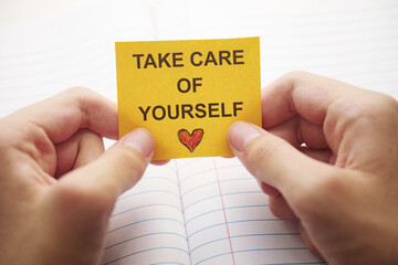 A young man holding a yellow paper note with the words Take care of yourself on it in his hands over an open notebook. Close up.