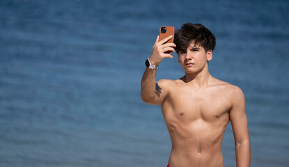 Shirtless young man taking a selfie with his smartphone on a sunny day with the ocean in the background with copy space