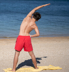 Rear view of teenager boy practicing side bending exercise on sandy beach with ocean in background