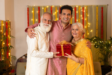 Indian happy young son giving red gift box to parents on the occasion of Diwali season in colorful...
