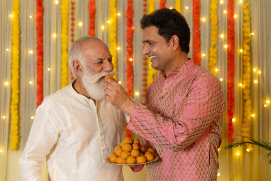 Indian smiling son feeding laddu to father while celebrating Diwali together on the occasion of Diwali season in colorful decorated home background - Diwali holidays festive