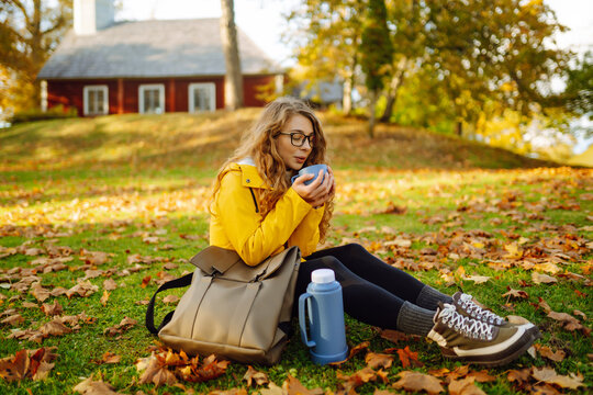 Female traveler in bright raincoat sits on a green meadow drinking hot tea from thermos surrounded by autumn foliage. Cute woman enjoys sunny autumn day in park. Weekend and adventure concept.