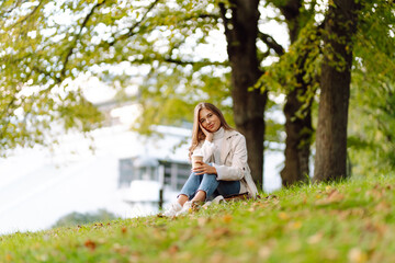 Obraz premium A beautiful woman sits on a blanket by a pond drinking a hot drink. Young woman posing in the park outdoors. Relaxation, nature concept.