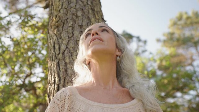 A serene mature woman with gray hair stands by a tree, embracing the tranquility of nature.