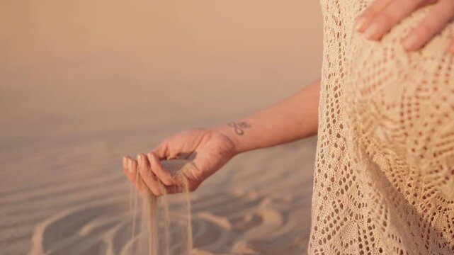 A pregnant woman stands in the desert at sunset. She holds her belly and gently lets sand fall from her hand.