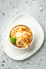 Hot French onion soup topped with melted cheese and croutons, garnished with parsley in a white bowl on a plate over a gray speckled surface, close-up, flat lay.