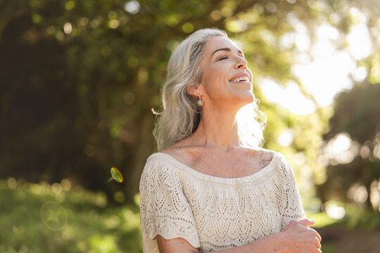 A joyful woman stands in a sunlit outdoor setting, embracing a peaceful moment. Her serene expression and the natural light highlight a sense of happiness and contentment.
