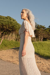 A graceful older woman with gray hair walks confidently through a lush, green outdoor setting. Her...