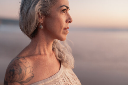 A mature woman with a tattoo gazes thoughtfully at the ocean during a calming sunset. The serene scene captures contemplation, tranquility, and the beauty of aging gracefully.