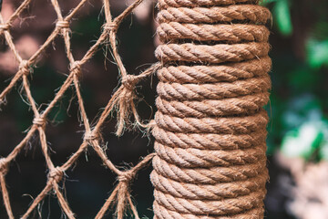 Thick rope coils tightly around a cylindrical post beside a knotted net, both showcasing coarse,...