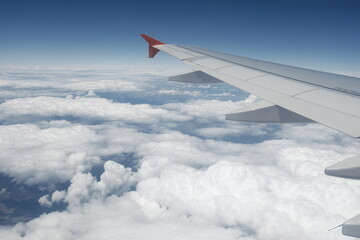 Fototapeta premium A peaceful in-flight view from a passenger seat showing an aircraft wing soaring above soft clouds under a bright blue sky.