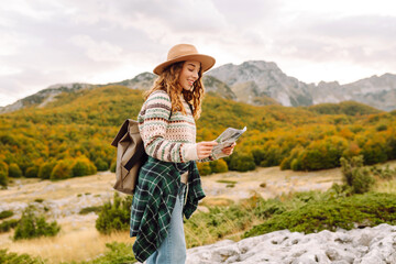 Fototapeta premium Smiling woman in backpack holding map against majestic mountains background. Young female traveler enjoying landscapes with map of area and choosing direction for hiking. Travel concept, navigation.
