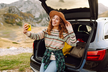 Traveler in car trunk with phone taking selfie or blogging against mountain landscape. Beautiful...