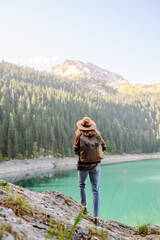A back view of a female hiker in a hat and with a backpack sitting on a rock overlooking a high mountain lake. Young woman feels freedom and enjoys mountain landscape. Hiking and nature concept.