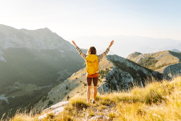 Cheerful woman with a bright hiking backpack walking on a picturesque mountain trail. Beautiful female traveler enjoying sunset in the mountains and feeling freedom. Hiking, freedom concept.