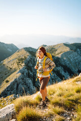 Naklejka premium Cheerful woman with a bright hiking backpack walking on a picturesque mountain trail. Beautiful female traveler enjoying sunset in the mountains and feeling freedom. Hiking, freedom concept.