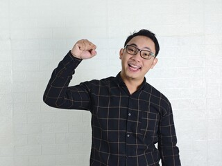 Energetic Asian Man with Glasses Celebrates Success Against White Brick Wall.