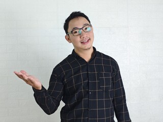 Smiling Asian Man Presenting with Open Palm Against White Brick Wall.