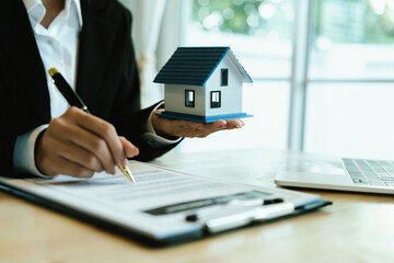 A real estate agent holds a house model and signs documents, symbolizing property sale, home purchase, financial agreements, mortgage loans, investment, and ownership in housing deals.