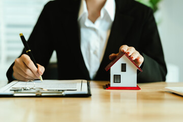 A real estate agent holds a house model and signs documents, symbolizing property sale, home...