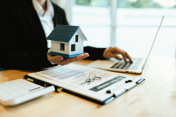 A businessperson signs a contract while holding a model house, symbolizing real estate investment,...