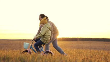 little child learns to ride bike with his mother, cheerful mother helps kid learn ride two-wheeled vehicle, baby pedals at sunset, daughter has fun in evening sun, girl with her mother ishappy family.