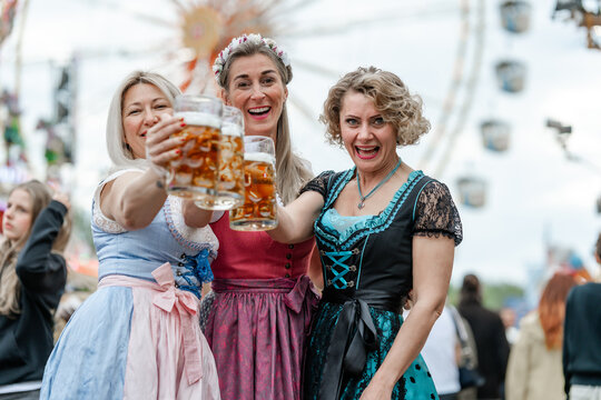 Three women in festive dirndl outfits clinking beer mugs during a sunny day at a traditional German beer festival