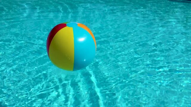 Vibrant colorful beach ball floating towards you in bright blue pool water with small waves and ripples of water on a bright sunny day. Pool toy floating in water