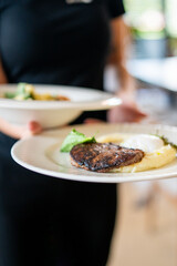 Server holding plate with grilled steak, mashed potatoes, poached egg, and greens in restaurant setting. Gourmet meal, close-up, handheld, indoor dining atmosphere.