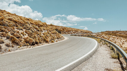 Curved road with guardrail in hilly landscape with arid vegetation under a blue sky with some clouds. Andalucia, Spain.