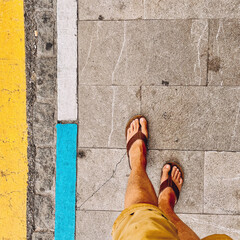 Tanned male feet in flip flops walking on pavement with yellow, blue and white marks. Seen from above.