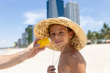 Father, applying sun cream on his  cute blond boy, playing on the sea, hat, sunglasses, swimsuit, sun protection cream, sea and sand background. Concept: love, lifestyle, children, vacations