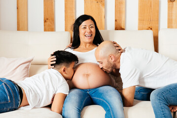 Eurasian Father and Son Joyfully Kiss Loving Pregnant Mother's Belly in Cozy Home Setting