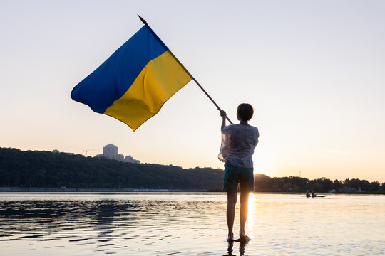 A woman waves a large yellow and blue Ukrainian flag fluttering at sunset while standing in the Dnipro River. Proud to be Ukrainian. Independence Day. Faith in victory. Support Ukraine. - Powered by Adobe