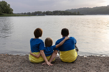 family of three in identical yellow and blue T-shirts, hugging, relax sitting on the bank of the Dnieper. National identity. Pride in being Ukrainian, faith in Ukraine's victory. Independence Day.