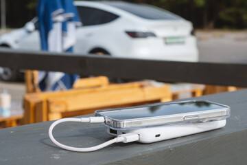 Smartphone connected to a portable power bank, charging outdoors on a wooden table. White electric...