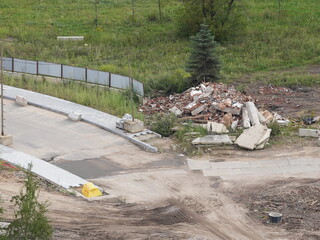 Urban demolition site with construction waste and debris near an unfinished road pavement and green field in background

