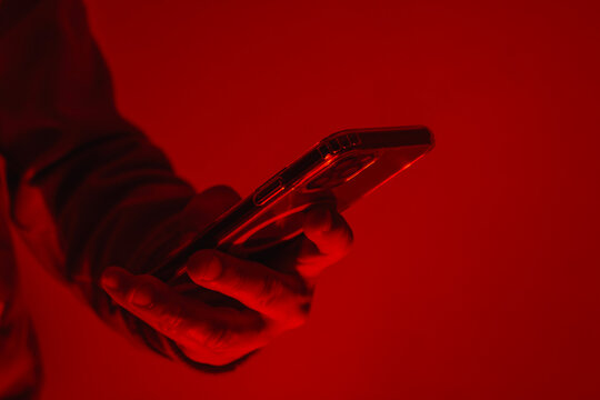 Close-up of a man's hand holding a modern smartphone under bright red studio lighting. Concept of digital cybersecurity, data privacy, hacker activity and darknet.