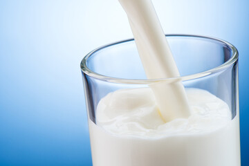 Fresh milk pouring into transparent glass on blue background